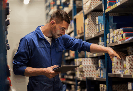 Worker in blue coveralls using tablet while pulling spare parts from rack - FORTNA